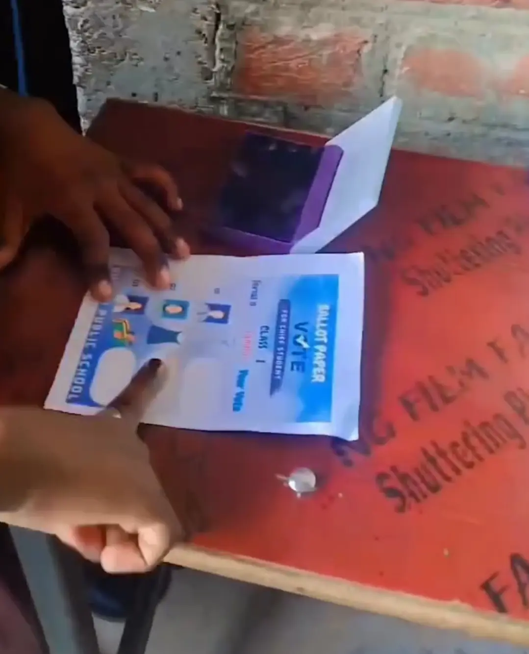 A child’s hands point to a Ruhani Public School ballot paper beside a purple ink pad.