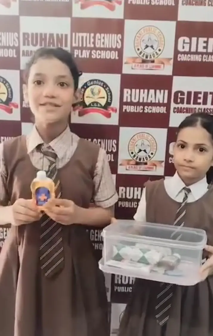 Two girls in school uniforms hold antiseptic and a first-aid kit in front of a school banner.