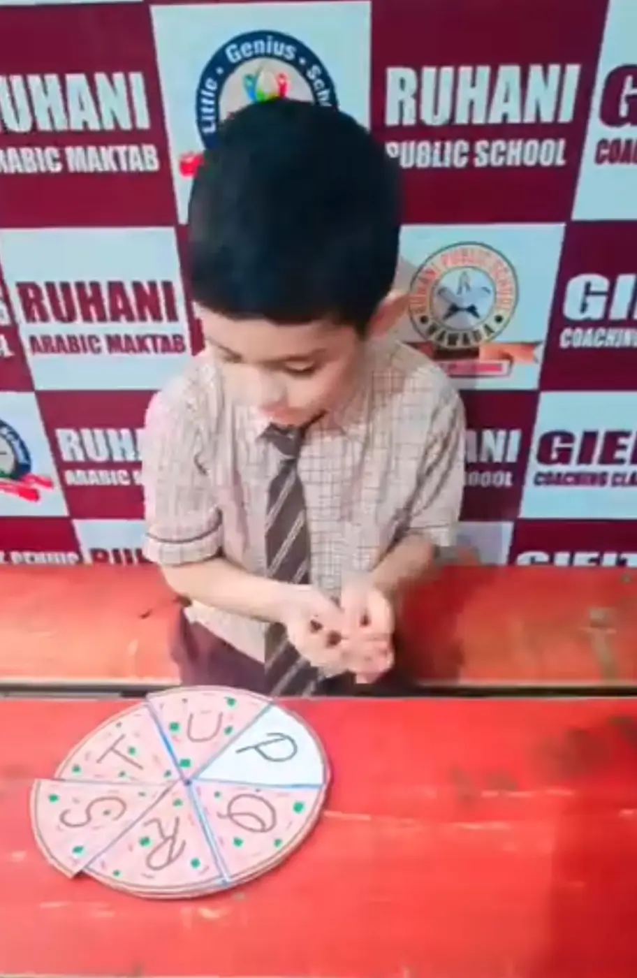A young boy studies a circular letter wheel with alphabet slices at a classroom desk.
