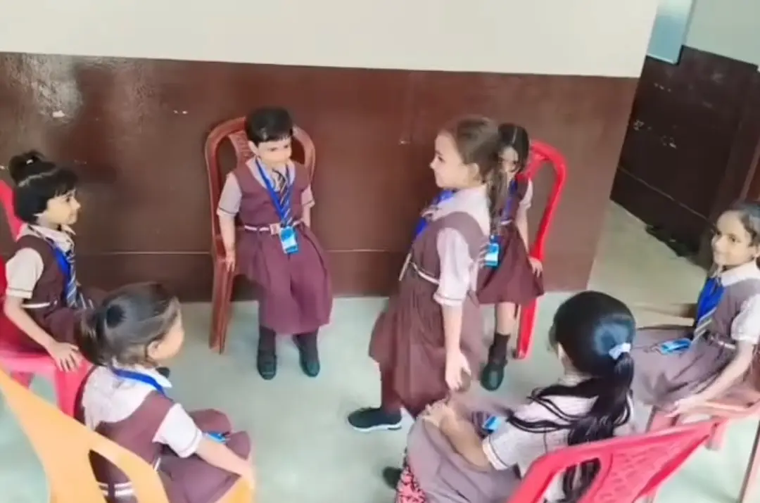 Young girls in brown uniforms sit in a circle of colorful chairs during class activity.