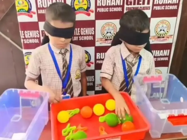 Two blindfolded boys reach into a red tray of colorful plastic balls and shapes.
