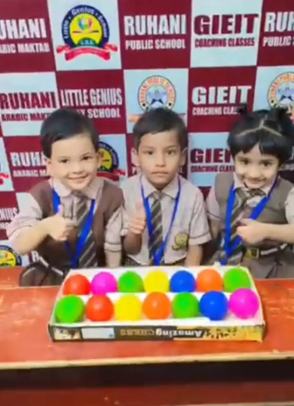 Three smiling students give thumbs up behind a box of colorful plastic balls.