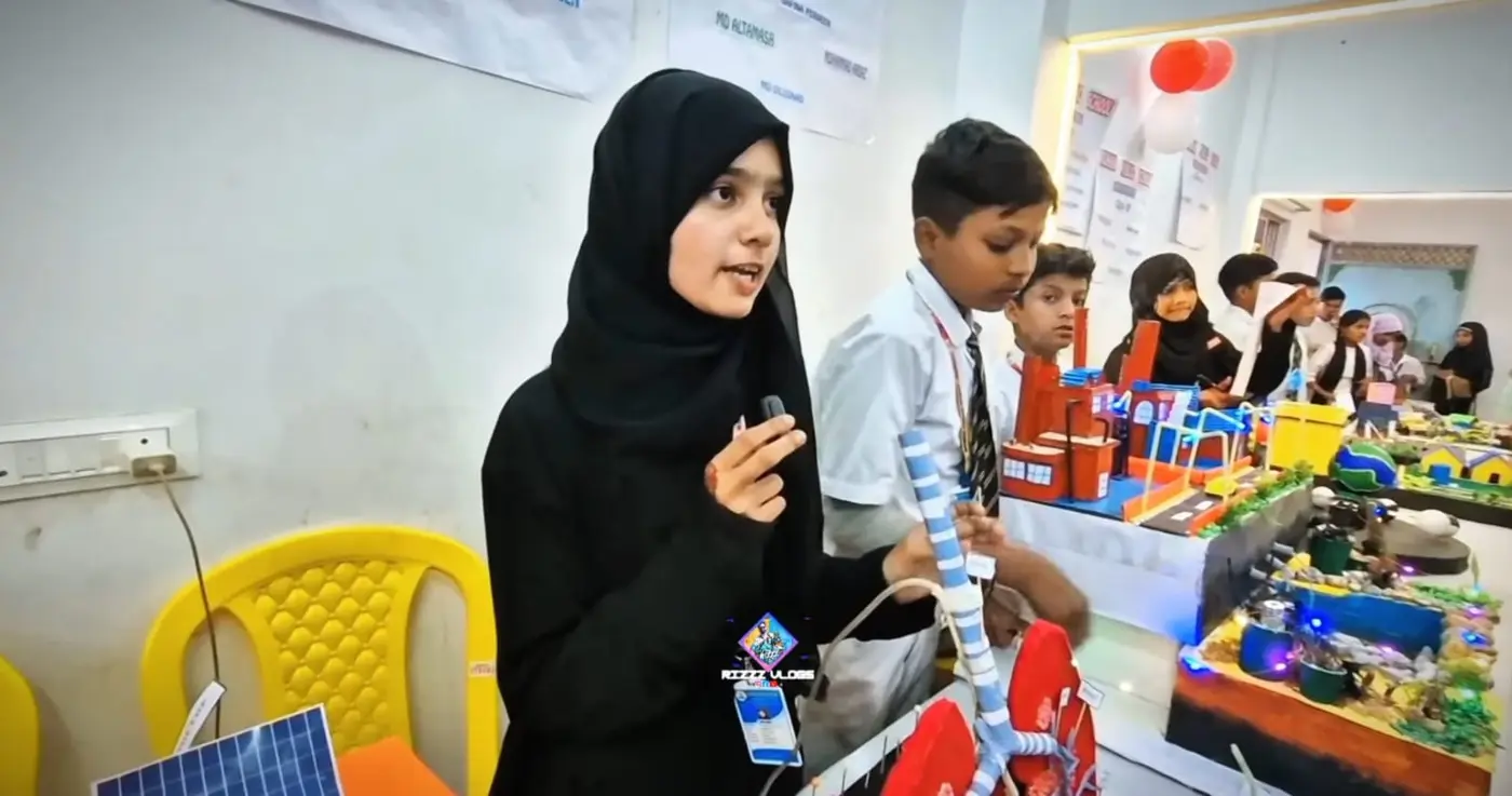 A student speaks into a microphone beside science project tables with colorful models.