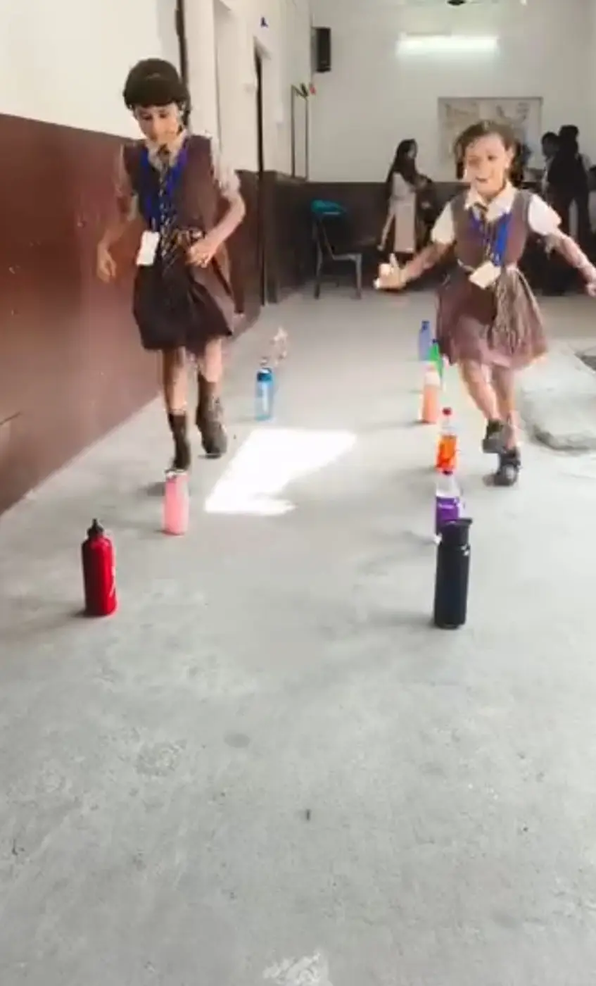Two girls in brown uniforms race past rows of water bottles used as hurdles.