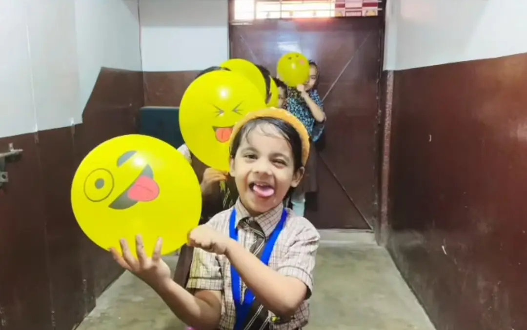 A smiling girl holds a yellow emoji balloon while classmates line up behind her.