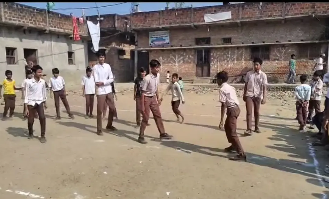 Boys in school uniforms play kabaddi on a chalk-marked dirt court beside school buildings.