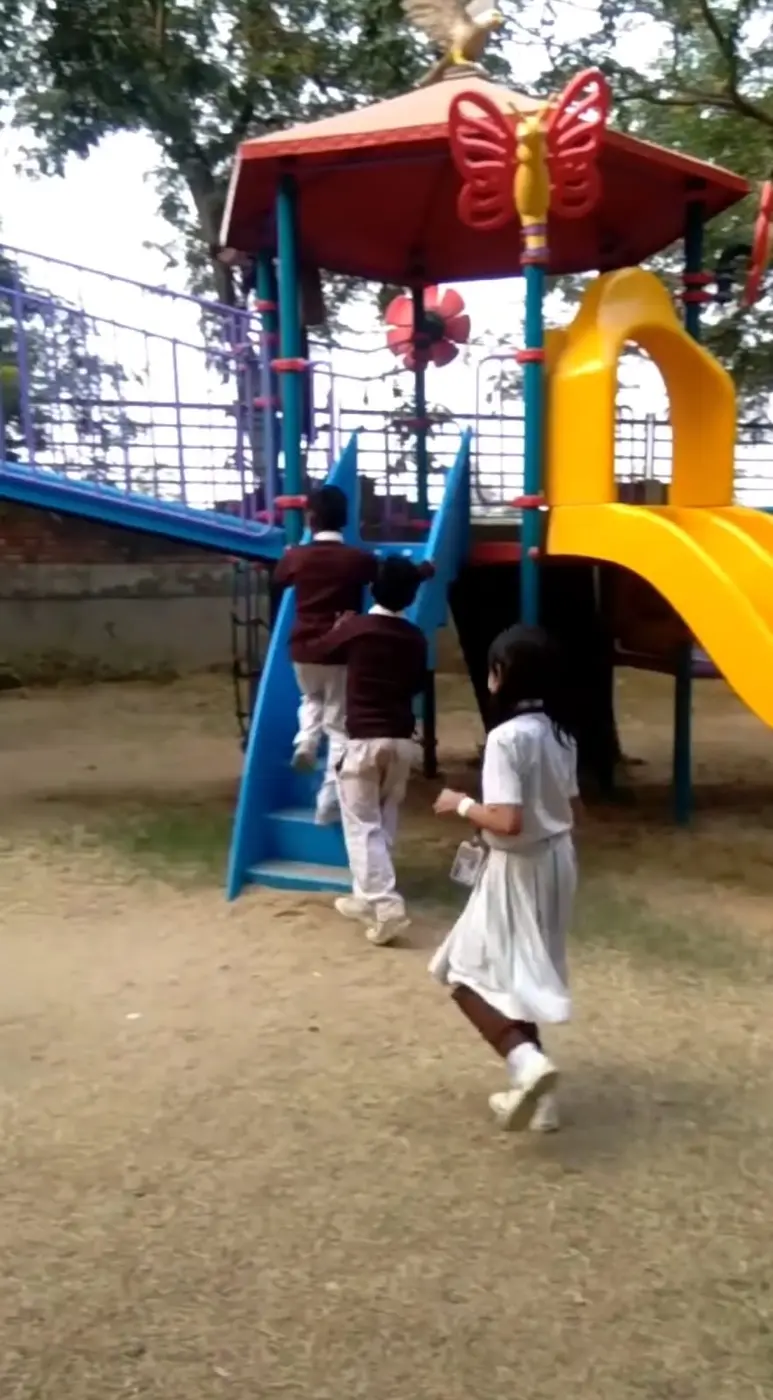 Students climb blue stairs on a red-roofed outdoor play structure with a yellow slide.
