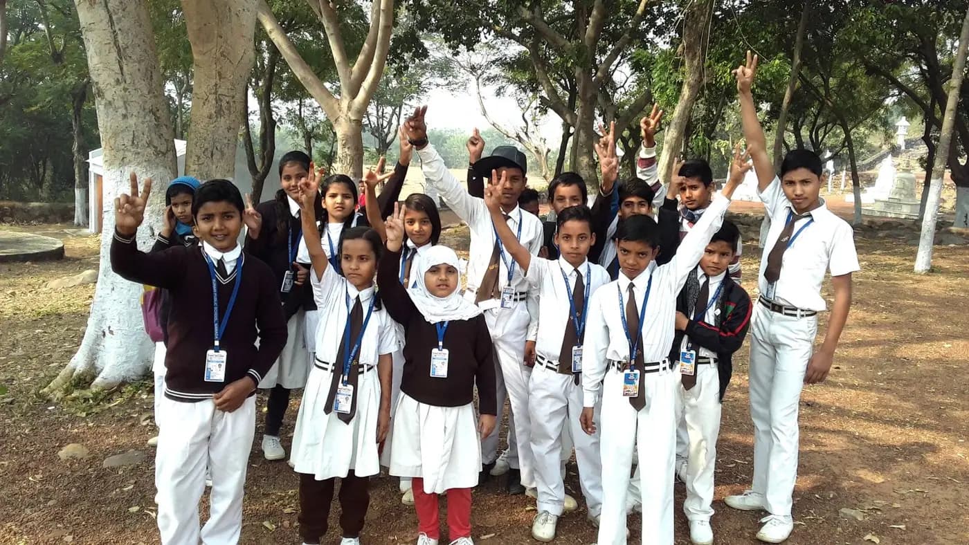 Students in white uniforms smile and make peace signs under shady trees.