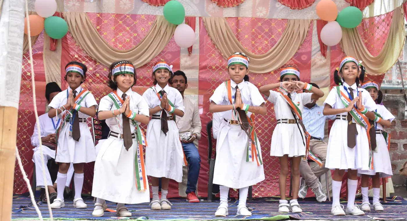 Girls in white uniforms and tricolor sashes stand in a namaste pose on stage.