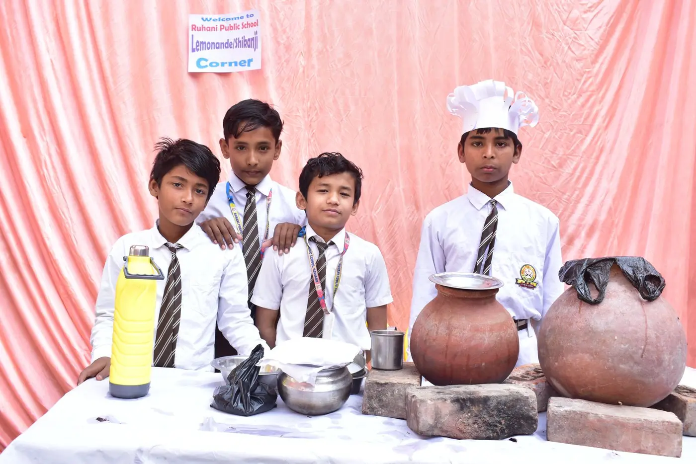 Boys in uniforms stand behind clay pots at a lemonade and shikanji stall.
