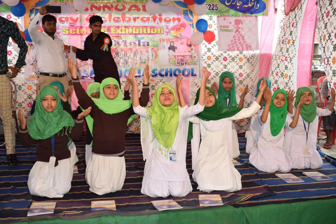 Girls in white outfits and green hijabs kneel with raised hands on the Annual Day stage.