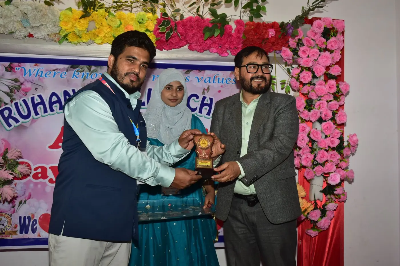 Guests present a wooden shield award on a flower-decorated Annual Day stage.