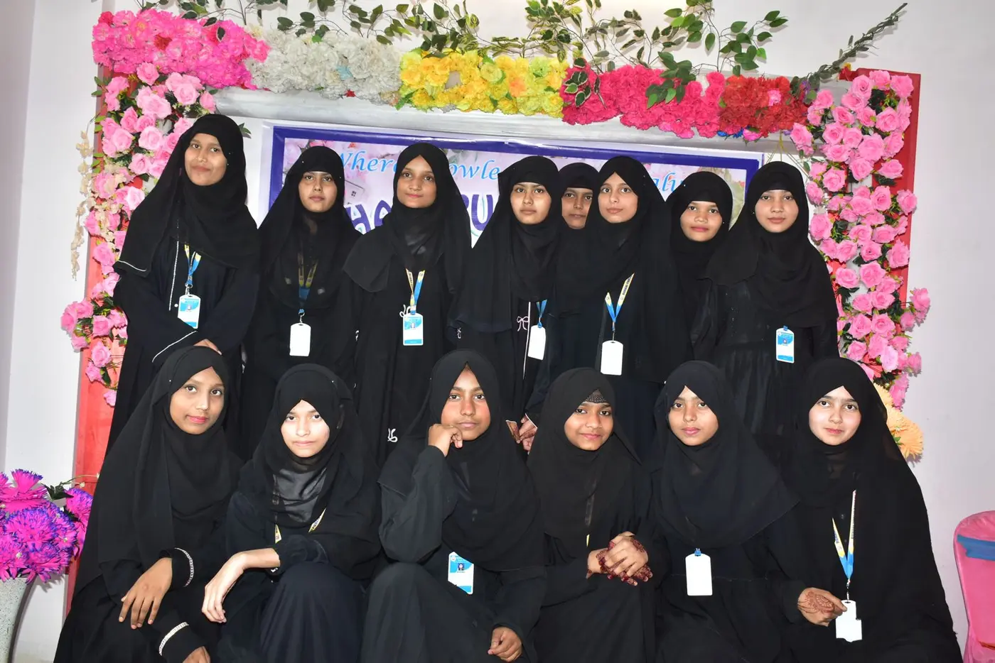 Students in black abayas and hijabs pose in two rows before a floral stage backdrop.