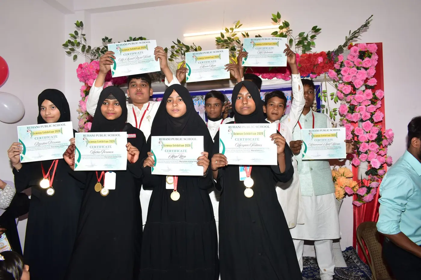 Students hold Science Exhibition certificates and medals in front of a floral arch.
