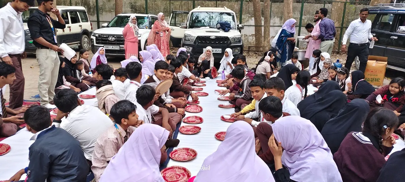 Rows of students in uniforms sit on white mats with plates during an outdoor communal meal.